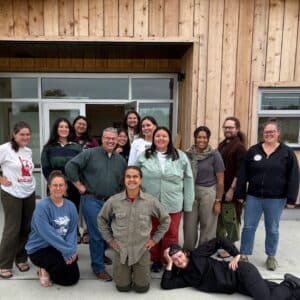 A group of people stand outside of a wooden building, posed and smiling at the camera.