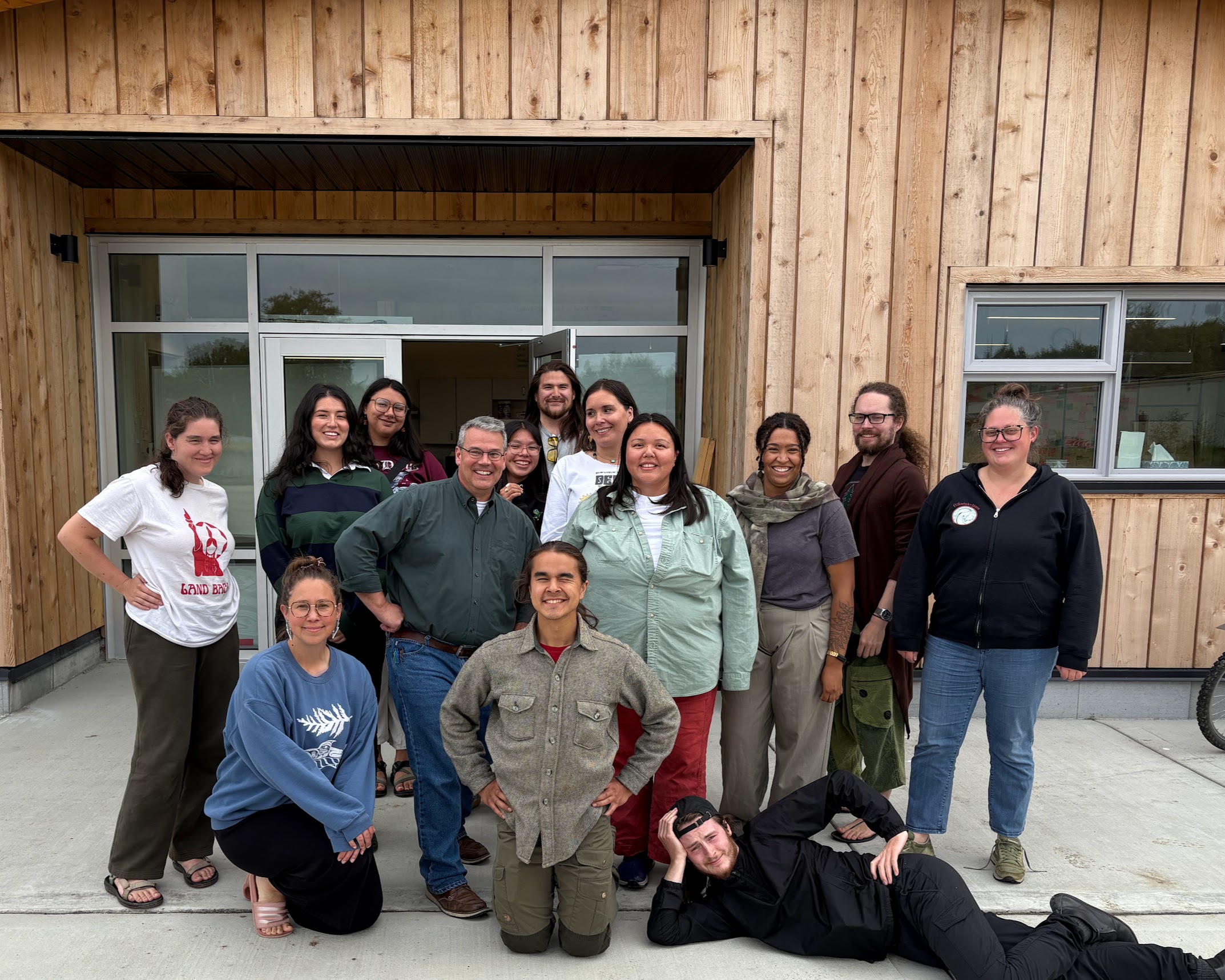 A group of people stand outside of a wooden building, posed and smiling at the camera.
