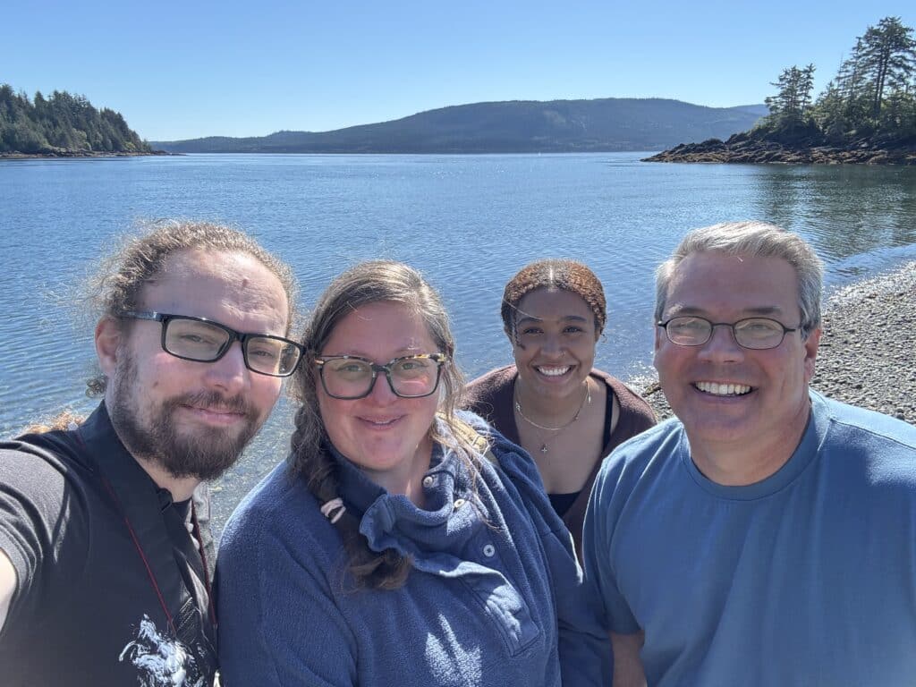 Cedar, Susanna, Reighen, and Evan stand shoulder to shoulder on a beach in the sun smiling at the camera 