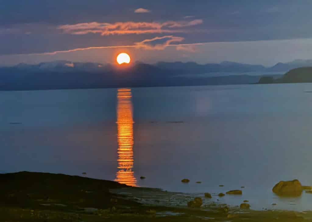 Bright orange sunset beyond the mountains over Hardy Bay with a trail of orange light across the flat water of the bay in the foreground.