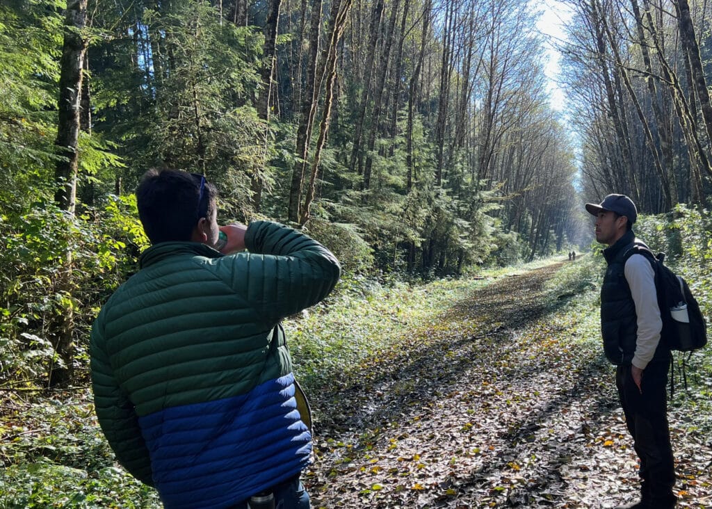 Zac and Steven on a Bak̕wa̱mk̓ala immersion language walk at the Quatse loop. Zac and Steven stand on a wide path with tall leafless trees standing tall on either side and shorter evergreen trees visible interspersed. Two other figures walk down the path in the far distance.