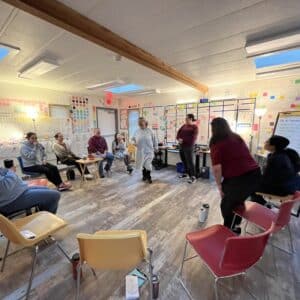 An immersion language session at the Treehouse in Port Hardy, BC. A circle of chairs fills the room while some students are standing and laughing. Colourful language posters and sticky notes are on the walls behind everyone.