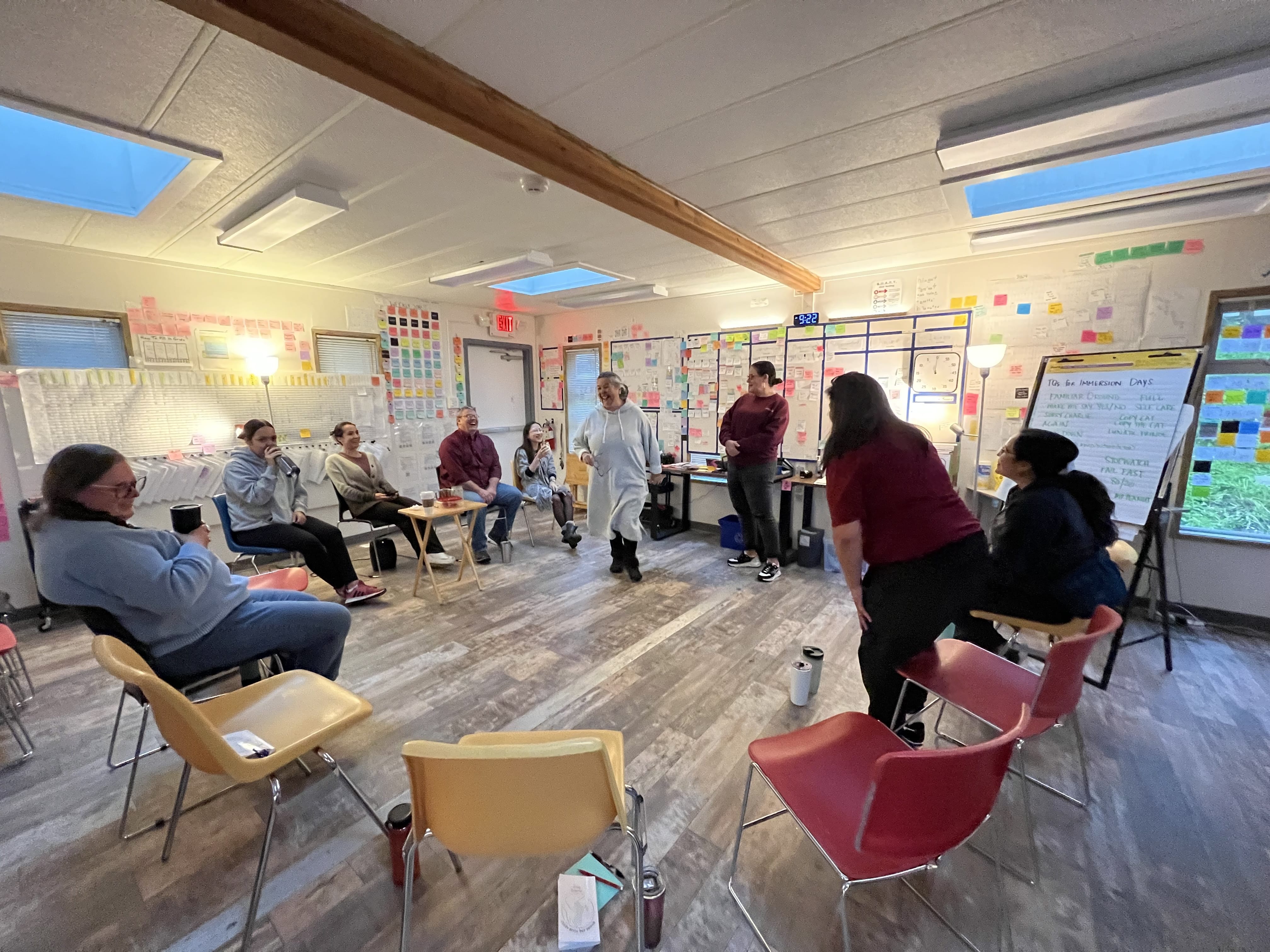 An immersion language session at the Treehouse in Port Hardy, BC. A circle of chairs fills the room while some students are standing and laughing. Colourful language posters and sticky notes are on the walls behind everyone.