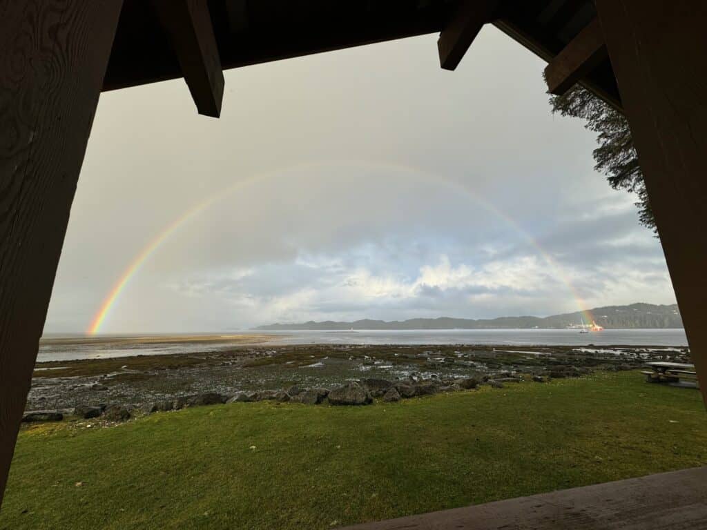 View of a rainbow over Hardy Bay in Port Hardy, BC with grass and a rocky beach at low tide in the foreground.